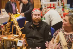 Stallholder wearing a festive Christmas hat chatting with visitors at a handmade wooden crafts stall